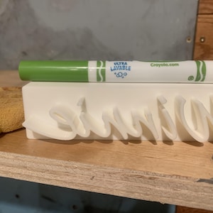 May include: A white 3D printed rectangular block featuring the raised cursive name Winnie on the front. A green Crayola Ultra Lavable marker rests horizontally on top of the block, and a copper penny sits to the left of the object.
