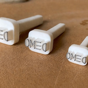 May include: Three white plastic stamps of varying sizes, each featuring the word DEMO in a clear, sans-serif font. The stamps have a rectangular base with rounded corners and a cylindrical handle extending from the back for easy gripping and use.
