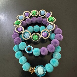 Op de afbeelding: Drie gestapelde armbanden met pluizige paarse en turquoise kralen. Eén armband bevat zilverkleurige ringen met kleine kleurrijke kralen. Een andere heeft een blauw-witte boze oog bedel. De derde bevat een goudkleurige ster en een gefacetteerde blauwe kraal.