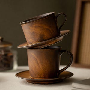May include: Two ceramic cups with matching saucers stacked vertically. The cups feature a dark brown, mottled, and textured finish with lighter amber-coloured streaks. Each cup has a curved handle and a smooth, wide rim, showcasing a rustic aesthetic.