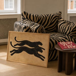May include: A rectangular framed print featuring two black panthers in a leaping motion against a tan background. The artwork is positioned in front of a zebra-striped armchair and next to a dark wood side table with a red book titled NEW YORK.