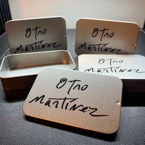 May include: A collection of rectangular metallic tins with rounded corners, featuring a matte finish. Each lid is printed with the black handwritten text Otro Martinez. One tin is shown open, revealing a hollow interior, while others are closed.