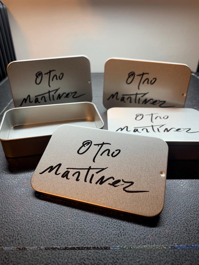 May include: A collection of rectangular metallic tins with rounded corners, featuring a matte finish. Each lid is printed with the black handwritten text Otro Martinez. One tin is shown open, revealing a hollow interior, while others are closed.