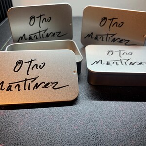 May include: A collection of rectangular silver metal tins with rounded corners. Each lid features the black printed text Otro Martinez in a stylized script font. Some tins are closed, while one is open, revealing the hollow interior space.