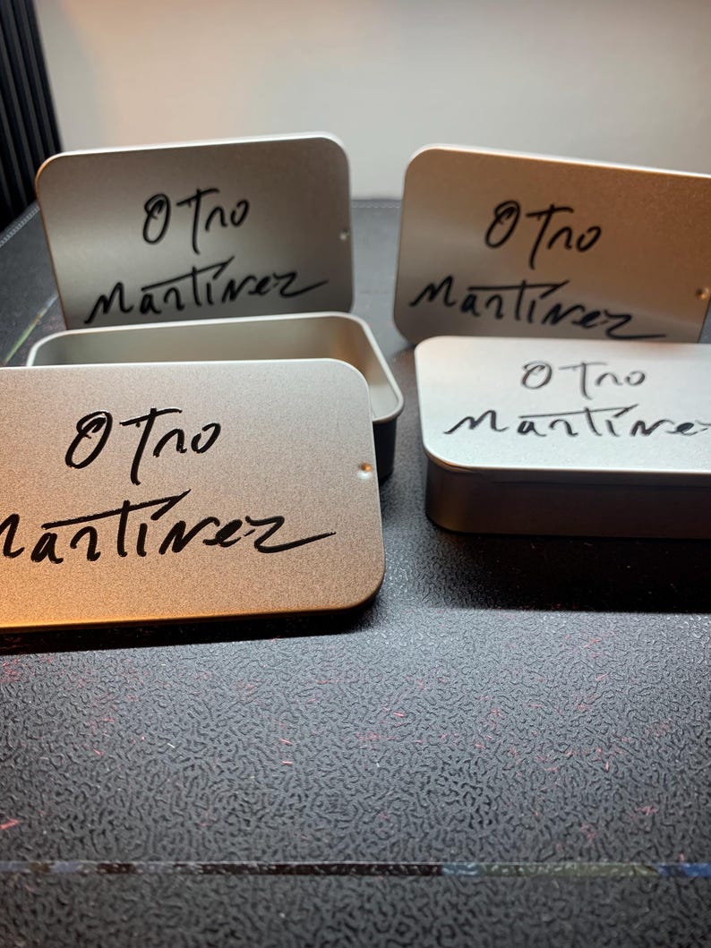 May include: A collection of rectangular silver metal tins with rounded corners. Each lid features the black printed text Otro Martinez in a stylized script font. Some tins are closed, while one is open, revealing the hollow interior space.