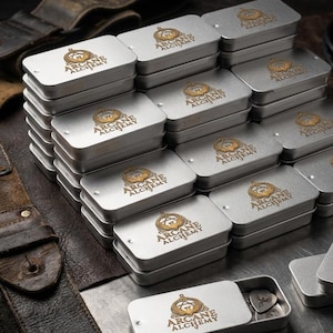 May include: A stack of rectangular silver metal tins, each featuring a gold-colored logo with the text Arcane Alchemy. One tin is open at the bottom, revealing a small guitar pick inside. The tins are arranged in a cluster on a dark surface.