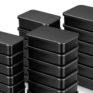 May include: A collection of rectangular black metal tins with lids, arranged in three separate stacks. Each individual container features a smooth matte black finish on the top and sides, with a thin silver-toned metallic rim visible at the base of each lid.