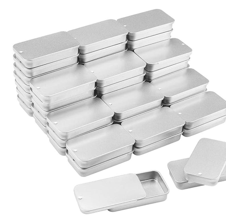 May include: A large collection of rectangular silver-colored metal tins with sliding lids. Each container features a smooth, metallic finish and a small circular indentation on the lid for easy opening. The tins are stacked in multiple rows.