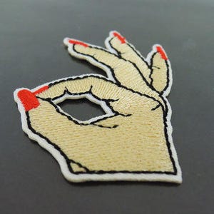 May include: A white embroidered patch of a hand making the OK sign with red nail polish.