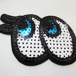 Op de afbeelding: Een zwarte en witte paillettenpatch met twee blauwe ogen. De patch is gevormd als twee ogen met wimpers.
