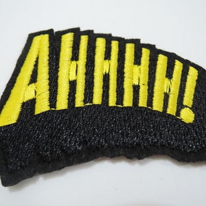 Puede incluir: Un parche de tela negro con letras bordadas en amarillo que deletrean "AHHHH!"