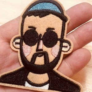 May include: Embroidered patch of a cartoon man wearing a blue hat, black sunglasses, and a black jacket. The man has a brown beard and mustache.