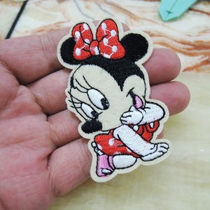 Könnte beinhalten: Ein gestickter Bügel-Patch mit einer Cartoon-Darstellung von Minnie Mouse mit einer roten Schleife und weißen Punkten. Sie trägt ein rotes Kleid mit weißen Punkten und weißen Schuhen.