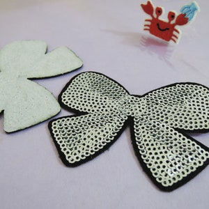 May include: Two iron-on patches in the shape of bows. One bow is white and the other is silver with sequins.