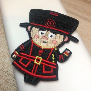 May include: Embroidered iron-on patch of a cartoon character wearing a black hat with a red band and a red and black uniform. The character has a smiling face with rosy cheeks.