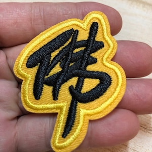 Puede incluir: Un parche bordado amarillo con costuras negras. El parche presenta el carácter chino para "fuerza" en una fuente estilizada. El parche mide 6 cm por 5 cm.