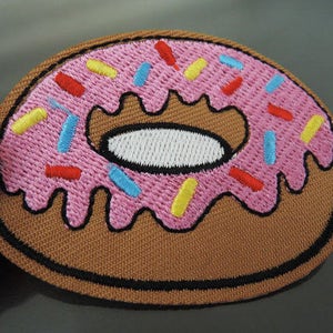 May include: An embroidered donut patch in pink and brown with colourful sprinkles. The donut has a white centre and is surrounded by a brown border.