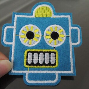 May include: Blue felt robot patch with white and yellow details. The robot has two large eyes with black pupils and white eyelashes. The robot has a mouth with white teeth and a yellow outline.