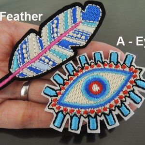 以下が含まれることがあります： 羽と目のデザインの刺繍パッチ。羽のパッチは、青、白、ピンクのディテールと黒い縁取りがあります。目のパッチは、青と赤のデザインと白い縁取りがあります。画像には「B - Feather」と「A - Eye」と書かれています。