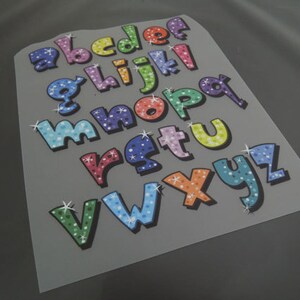 May include: A sheet of colorful iron-on letters with a sparkly finish. The letters are in a playful font and are perfect for adding a personal touch to clothing or crafts.