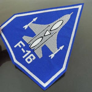 May include: A blue and white embroidered patch featuring a fighter jet with the text "F-16" in white.