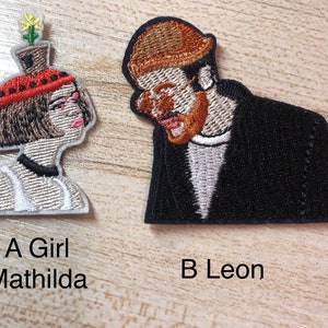 May include: Two embroidered iron-on patches, one of a girl with a flower on her head and the text "A Girl Mathilda" below, and the other of a man with a hat and the text "B Leon" below.