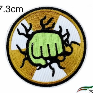 Könnte beinhalten: Ein runder gestickter Aufnäher mit einem Durchmesser von 7,3 cm, weißem Hintergrund und goldener Umrandung. In der Mitte des Aufnähers befindet sich eine grüne Faust mit schwarzen Umrissen und schwarzen Ranken, die sich von der Faust erstrecken. Die Ranken sind von einem goldenen und weißen Strahlungssymbol umgeben.