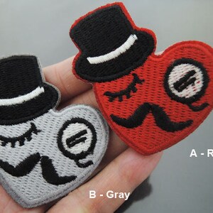 May include: Two iron-on patches in the shape of a heart with a top hat and a mustache. The patch on the left is gray with a black top hat and mustache. The patch on the right is red with a black top hat and mustache. The patches are labeled A - Red and B - Gray.