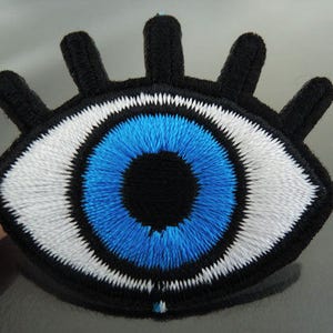 Può includere: Un cerotto per l'occhio ricamato in bianco e nero con un'iride blu e una pupilla bianca. Il cerotto per l'occhio ha una frangia attorno ai bordi.