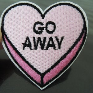 May include: Embroidered heart-shaped patch in light pink with the words "GO AWAY" in black. The patch has a white border and a pink stripe across the bottom. The patch is likely for clothing or accessories.