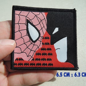 Könnte beinhalten: Ein roter und schwarzer gestickter Aufnäher mit einer Nahaufnahme von Spider-Mans Gesicht links und Batmans Gesicht rechts. Der Aufnäher ist 6,5 cm x 6,3 cm groß.