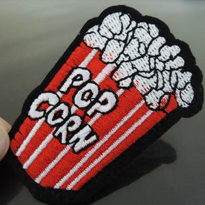 Może przedstawiać: Naszywka haftowana na czerwono i biało z napisem "Popcorn" w kolorze czarnym. Naszywka przedstawia kreskówkową wersję czerwono-białego wiaderka z popcornu, z którego wysypuje się popcorn.