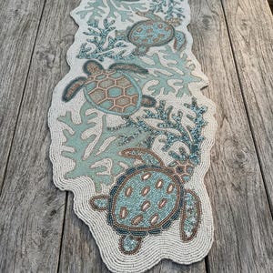 May include: A beaded table runner featuring a white background with a repeating pattern of sea turtles and coral branches in shades of light blue, teal, and metallic bronze. The runner has an irregular, organic shape with a textured beaded surface.