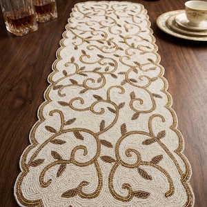 May include: A rectangular table runner featuring a scalloped edge and an intricate beaded design. The runner is composed of white beads forming the background with gold beads creating a swirling vine and leaf pattern across the surface.