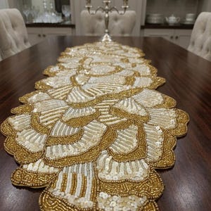 Puede incluir: Un camino de mesa largo y estrecho con un diseño intrincado de bordes festoneados. La superficie está densamente adornada con cuentas de vidrio doradas y blancas y lentejuelas circulares planas, dispuestas en un patrón repetitivo de abanicos superpuestos.