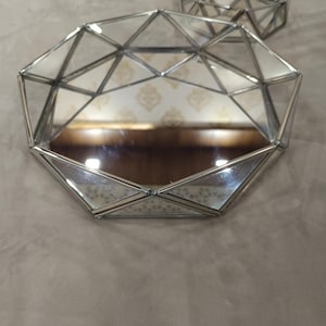 Op de afbeelding: Twee achthoekige dienbladen met spiegelende bodems en zilverkleurige metalen frames. De zijkanten zijn opgebouwd uit heldere glaspanelen die bij elkaar worden gehouden door dun soldeerwerk, wat een geometrisch, gefacetteerd uiterlijk geeft.