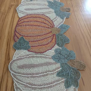 Può includere: Un runner da tavola lungo e stretto con un design a perline che raffigura quattro zucche nei toni dell'arancione, crema e beige. Le zucche sono arricchite da foglie di perline verdi e steli dorati, realizzati con intricate perline.