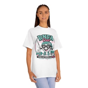 May include: A white t-shirt featuring a graphic of a cartoon character wearing a teal surgical cap and sunglasses. The text reads RNFA Crew, Scrub Skill, Focus Care, and Behind Every Surgeon Is An RNFA, Precision, Partnership, Pride.
