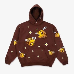 May include: A dark brown hooded sweatshirt featuring a repeating pattern of yellow and black pixelated bees and small white cross-shaped icons. The garment includes a front kangaroo pocket, long sleeves, and a hood with a single white cross detail.