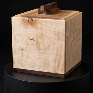 May include: A square wooden box featuring light-colored wood grain on the sides and top, accented by a dark brown wood base and a small, rectangular dark brown wood handle centered on the lid. The box displays natural wood knots and patterns.
