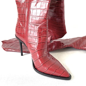 Puede incluir: Botas rojas de cuero con estampado de cocodrilo, por encima de la rodilla, con punta puntiaguda y tacón de aguja.