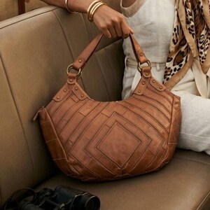May include: A brown leather hobo bag featuring a geometric patchwork design with visible stitching, gold-tone metal hardware, and two sturdy shoulder straps. The bag has a structured, curved silhouette and a textured finish on the leather panels.