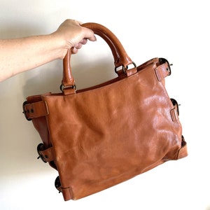 May include: A brown leather tote bag with two handles and multiple decorative buckles. The bag is held by a person's hand. The bag has a rectangular shape and a worn appearance.