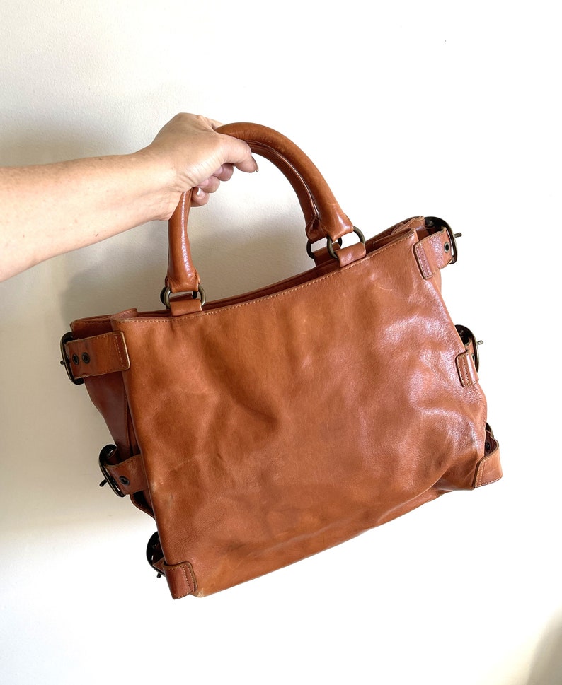 May include: A brown leather tote bag with two handles and multiple decorative buckles. The bag is held by a person's hand. The bag has a rectangular shape and a worn appearance.