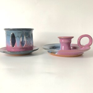 May include: Two ceramic candle holders. One is a cylinder with oval cutouts, the other a saucer with a handle. Both are glazed in blue, pink, and purple hues, with a rustic, handmade appearance.