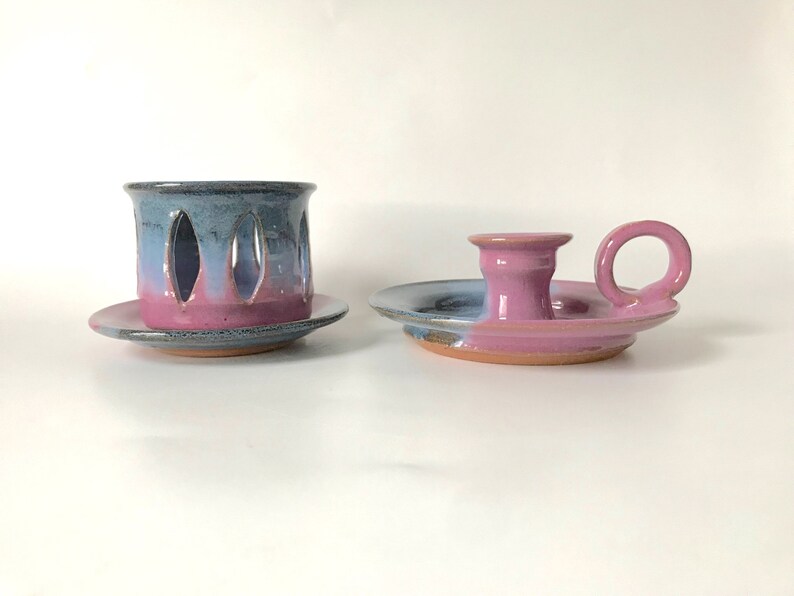 May include: Two ceramic candle holders. One is a cylinder with oval cutouts, the other a saucer with a handle. Both are glazed in blue, pink, and purple hues, with a rustic, handmade appearance.