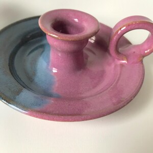 May include: A handcrafted ceramic candle holder with a circular base and a handle. The candle holder is primarily pink with a blue and purple gradient on the base. The handle is a loop shape, and the candle holder has a glossy finish.