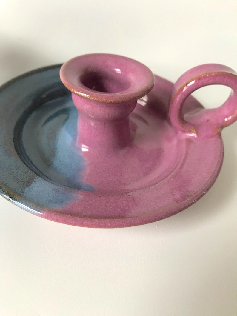 May include: A handcrafted ceramic candle holder with a circular base and a handle. The candle holder is primarily pink with a blue and purple gradient on the base. The handle is a loop shape, and the candle holder has a glossy finish.