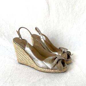 May include: A pair of metallic gold wedge sandals with a bow detail on the toe. The sandals have a woven espadrille wedge heel and adjustable ankle straps. The shoes are displayed on a white background.