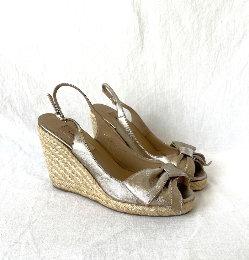 May include: A pair of metallic gold wedge sandals with a bow detail on the toe. The sandals have a woven espadrille wedge heel and adjustable ankle straps. The shoes are displayed on a white background.